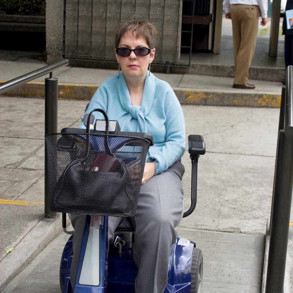 A woman using a mobility scooter on a concrete sidewalk ramp with handrails, illustrating ADA-compliant sidewalk access in a California commercial property setting