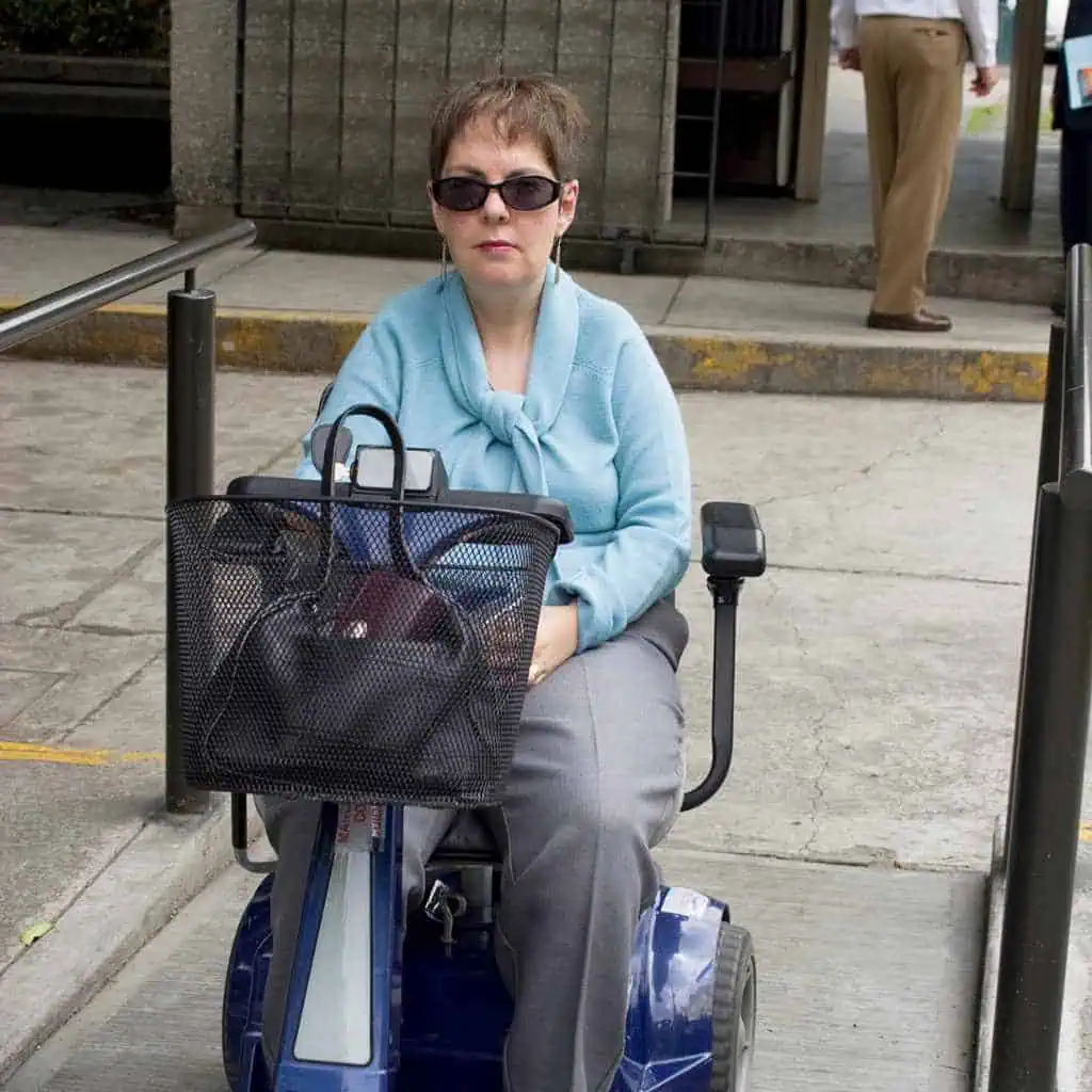 A woman using a mobility scooter on a concrete sidewalk ramp with handrails, illustrating ADA-compliant sidewalk access in a California commercial property setting