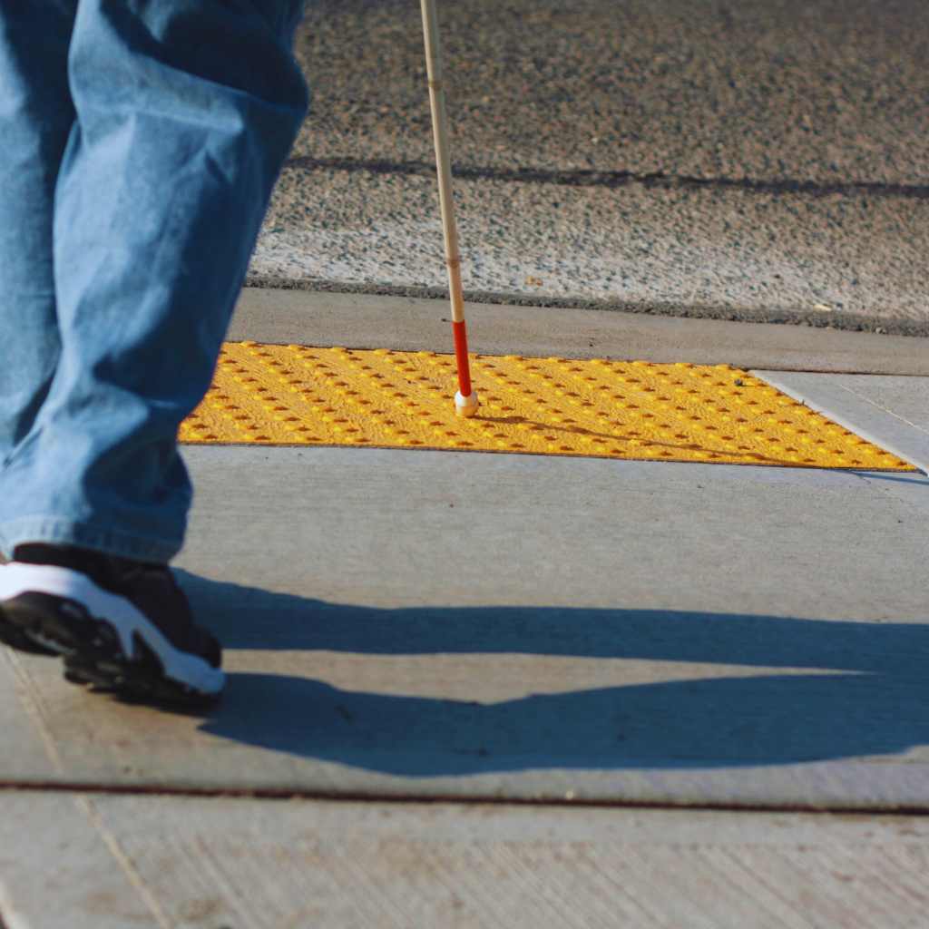 A person using a white cane steps onto a bright yellow tactile warning surface at a sidewalk curb ramp, illustrating ADA-compliant design for visually impaired access in California