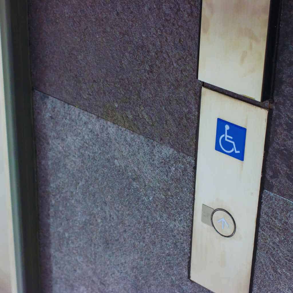 An elevator hall call button mounted beside the doorway with the blue wheelchair accessibility symbol, showing required identification and accessible reach height under ADA and California Title 24.