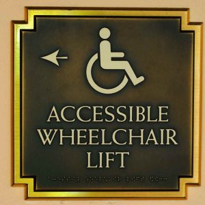 Bronze accessibility sign with wheelchair symbol and Braille, indicating the location of an accessible lift in a California building.