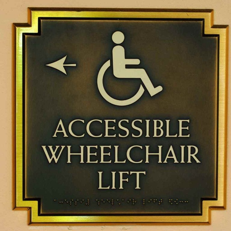 Bronze accessibility sign with wheelchair symbol and Braille, indicating the location of an accessible lift in a California building.