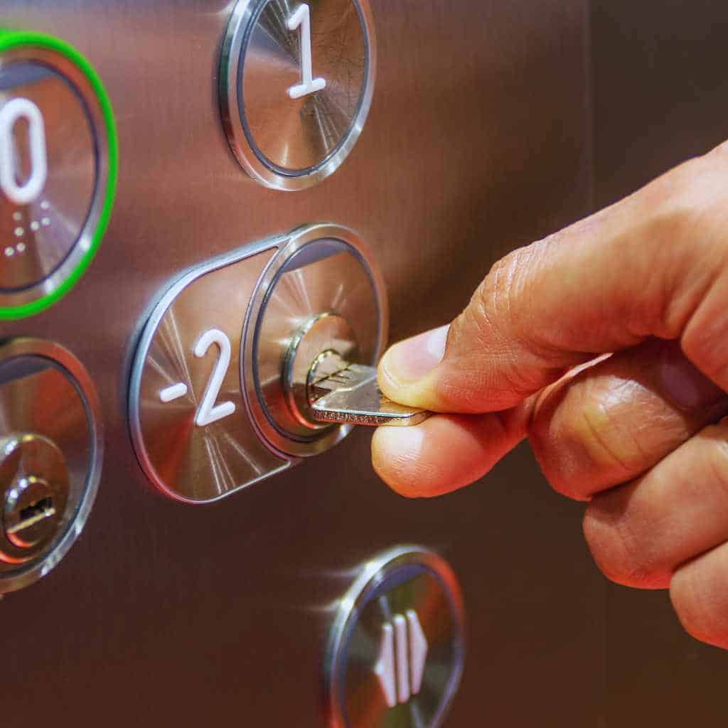 A person using a key switch on an elevator control panel, showing raised floor buttons and illuminated indicators that relate to ADA and California Title 24 accessibility requirements for reach range, tactile features, and operable parts.