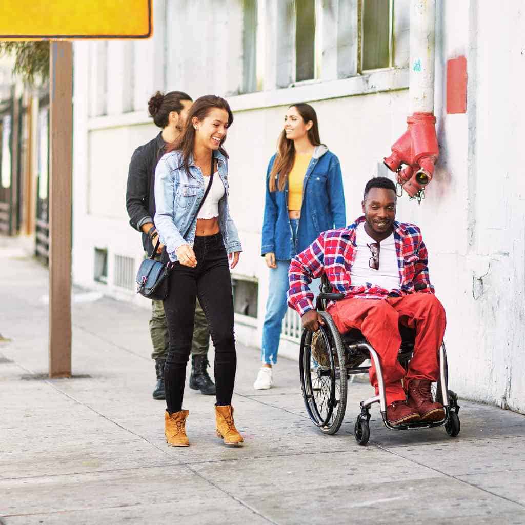 A man using a wheelchair and a group of young adults walking along a wide, unobstructed city sidewalk—illustrating inclusive, ADA-compliant pedestrian infrastructure in California.