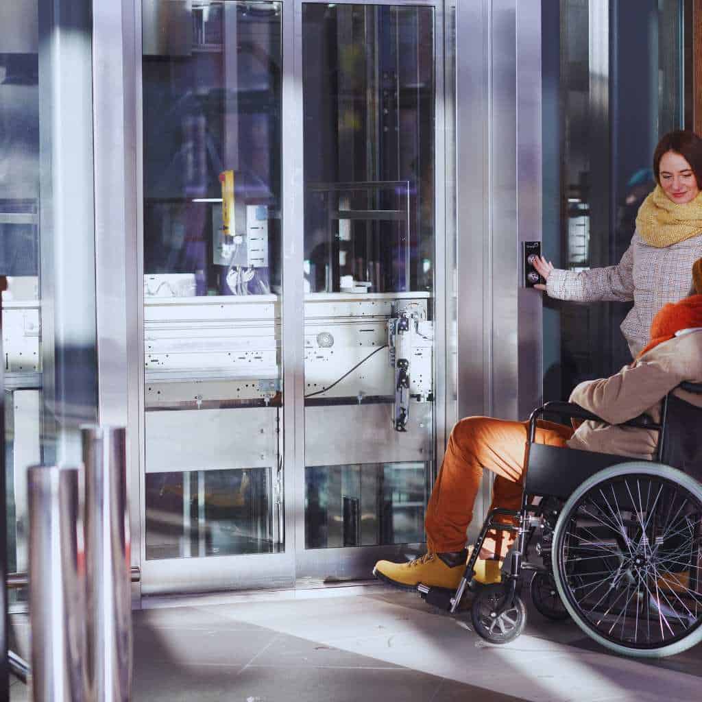 Person in a wheelchair entering a glass elevator while another user presses the hall call button, showing accessible operation features required under ADA and California Title 24.