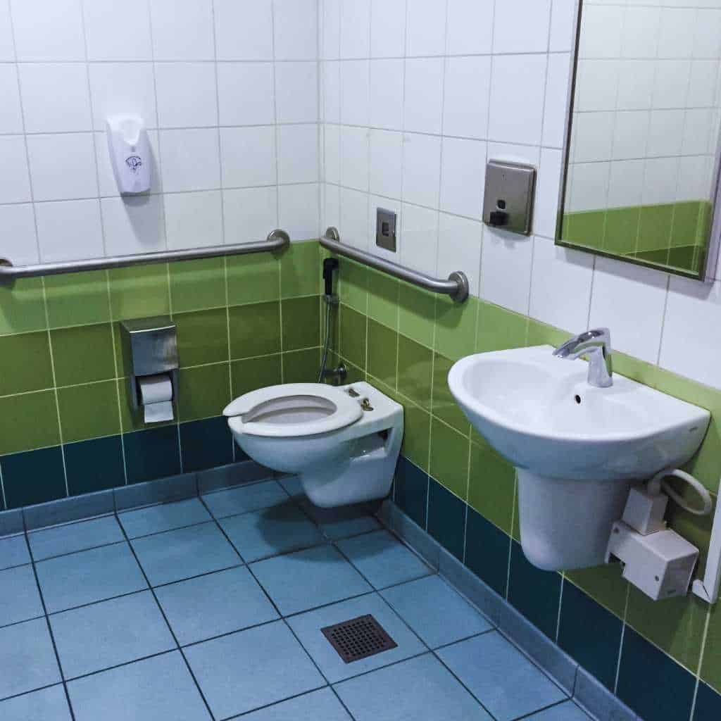 Restroom with wall-mounted toilet, side and rear grab bars, accessible sink, and clear floor space showing a typical ADA and Title 24 compliant fixture layout.