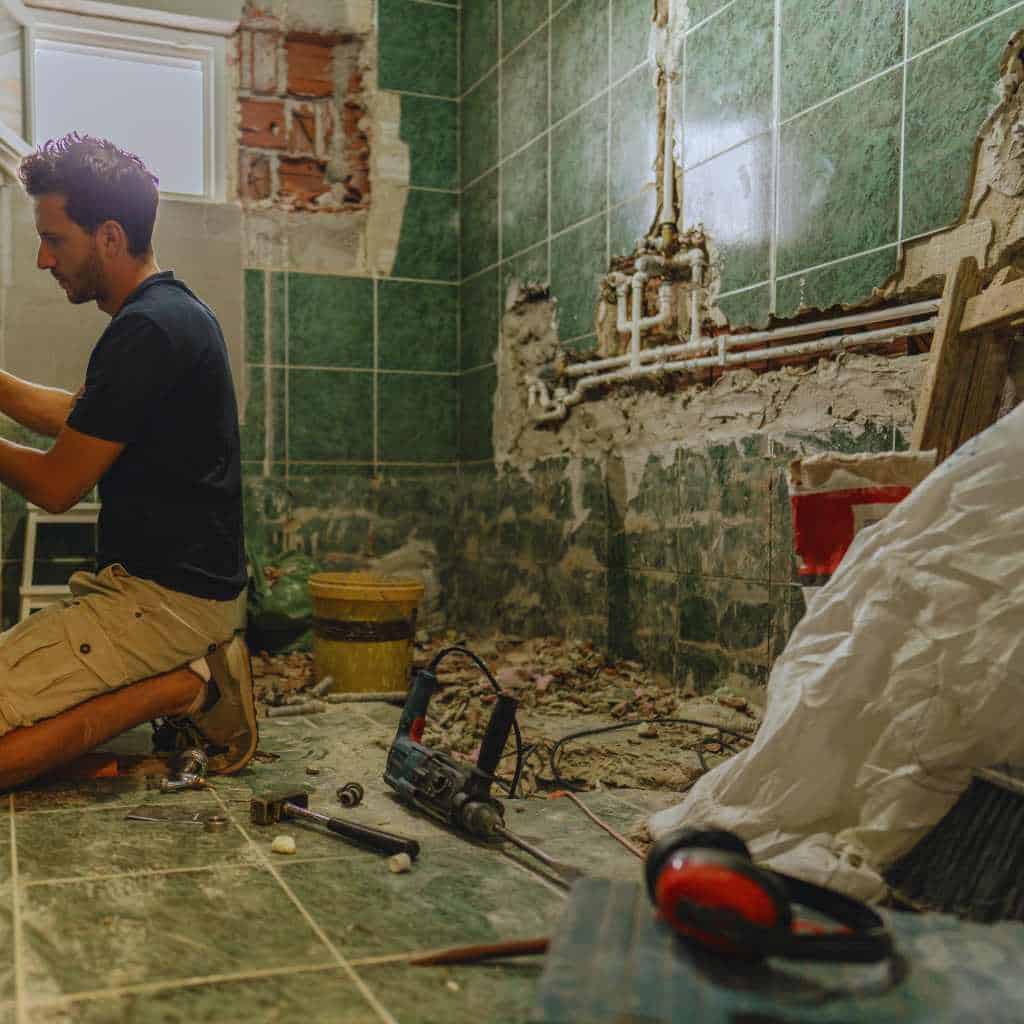 Contractor working inside a partially demolished bathroom during renovation, with exposed plumbing and tools on the floor, showing conditions before ADA and Title 24–compliant fixtures and clearances are installed.