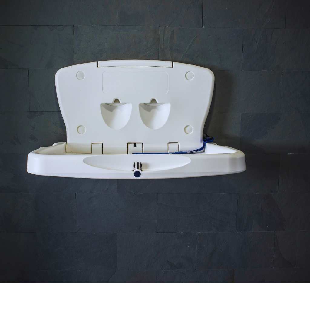 A white horizontal baby changing table securely mounted to a dark slate wall, featuring dual compartments and a safety belt.