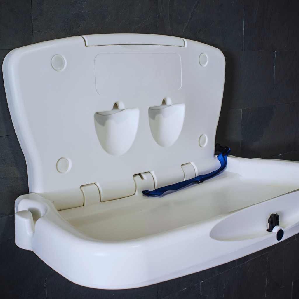 ADA-compliant baby changing station mounted on a dark tiled wall, shown in open position with visible safety strap and molded compartments for storage