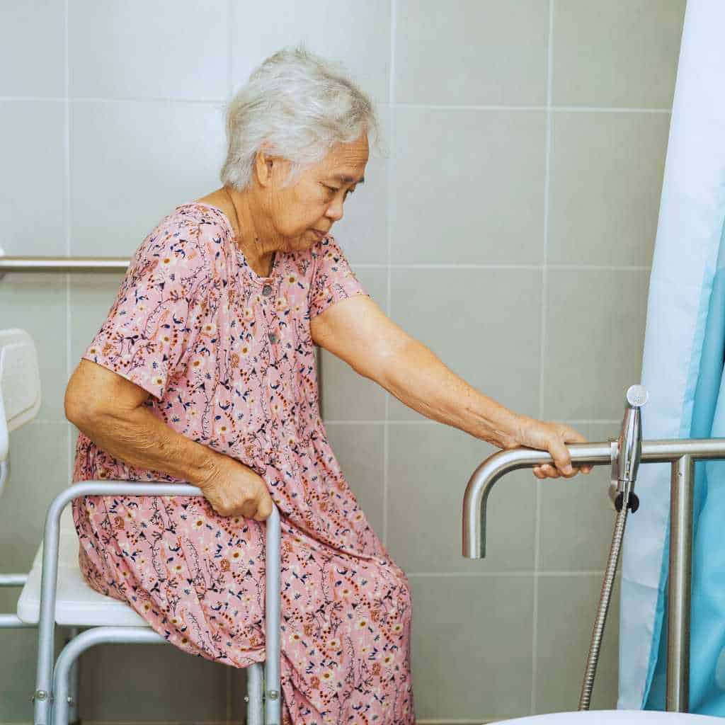 Older woman seated on a shower chair using ADA-style grab bars to stabilise herself while moving in a tiled bathroom, illustrating safe mobility support for users with limited strength or balance.