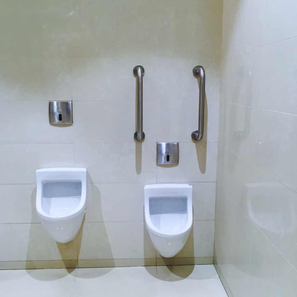 Two wall-mounted urinals with vertical and horizontal grab bars installed to support users, showing an accessibility layout often required in California public restrooms.
