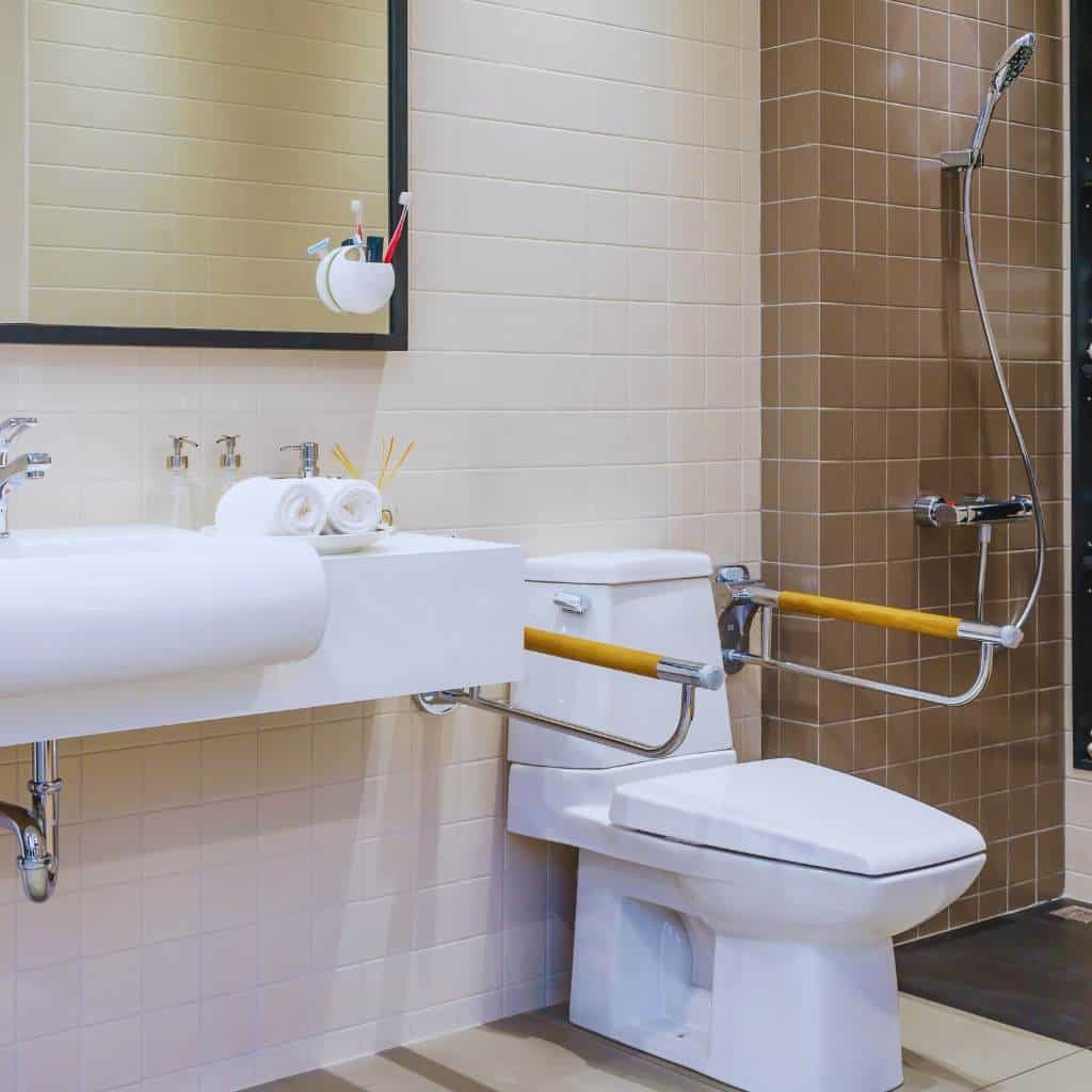 Accessible bathroom showing a toilet with side and rear grab bars, an open sink with knee clearance, and a walk-in shower with handheld spray and control valve
